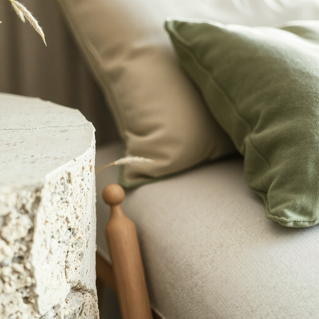 A close-up detail shot focusing on the juxtaposition of materials, showing a rough travertine side table beside a soft velvet cushion in sage green, capturing the tactile contrast and earthy elegance essential to modern organic design under soft natural light.